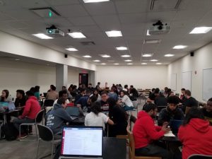 A meeting room where dozens of students sit, grouped at tables, talking and working on laptops.